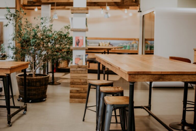 Mesa larga de madera con taburetes metálicos en un ambiente de oficina con plantas y decoración cálida