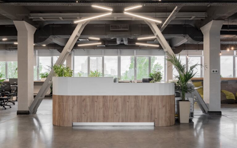 Mostrador de recepción curvado con acabado de madera y blanco, rodeado de plantas y elementos industriales en un espacio de oficina amplio y luminoso.